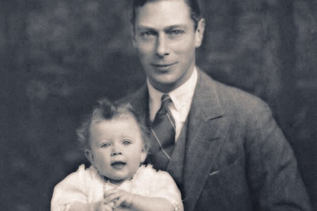 King George VI holds a baby Queen Elizabeth on his knee in a black and white photograph.