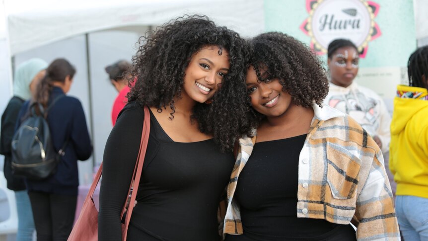 Two very attractive black women with curly hair, both smiling and wearing black tops, one with a tartan coat