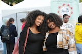 Two very attractive black women with curly hair, both smiling and wearing black tops, one with a tartan coat