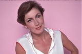 A woman wears a white top and red skirt in front of a pink background.