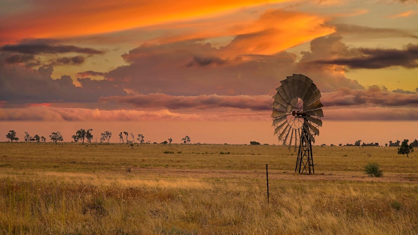 A short windmill standing in a country paddock, with the sun setting in the sky.