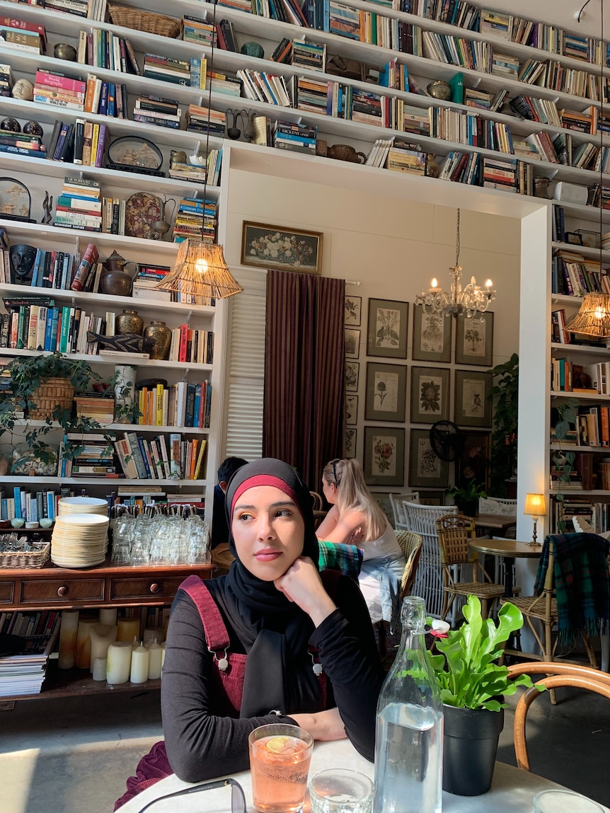 A young woman in a hijab sits at a table in a cafe surrounded by shelves filled with books.