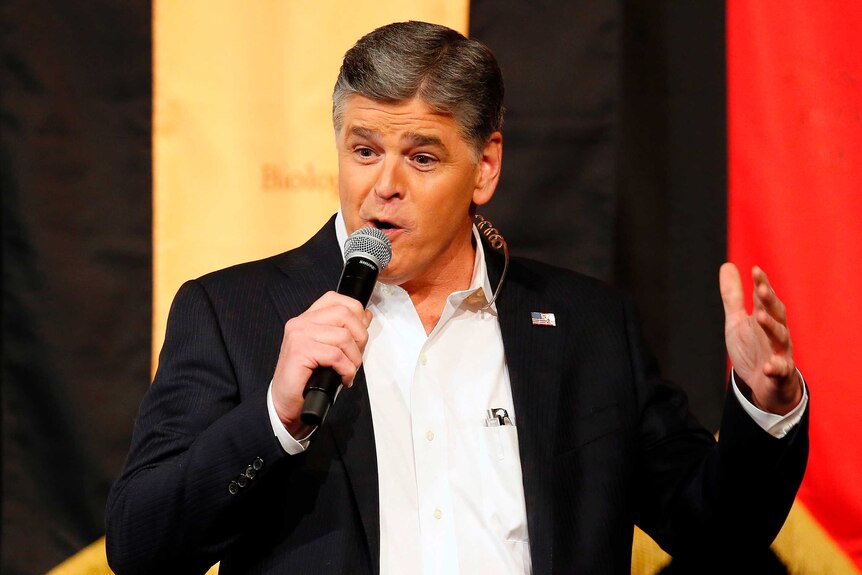 Sean Hannity holds a microphone and speaks, in front of a coloured banner.