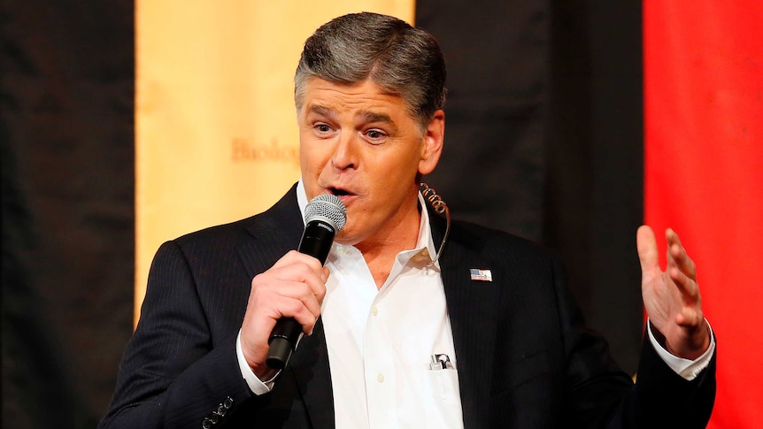 Sean Hannity holds a microphone and speaks, in front of a coloured banner.