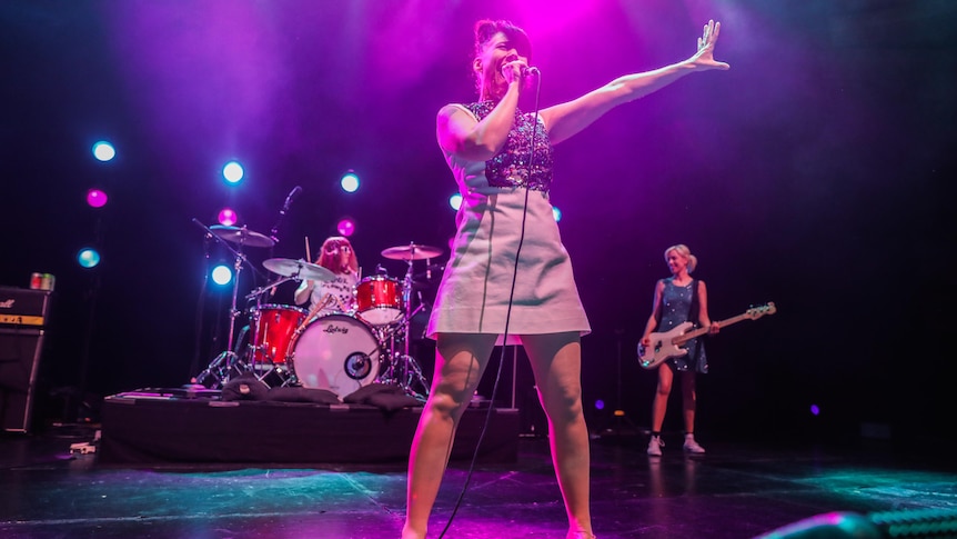 Three members of Bikini Kill performing on a big stage. Kathleen Hanna has an arm stretched out and holds microphone