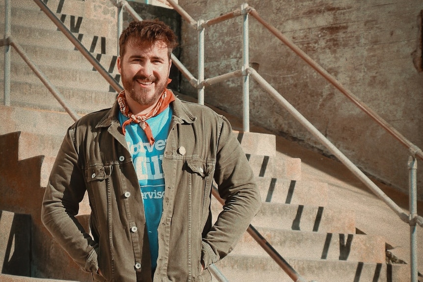 A man wearing blue Tshirt, khaki jacket and red neckerchief stands in front of cement stairs, smiling