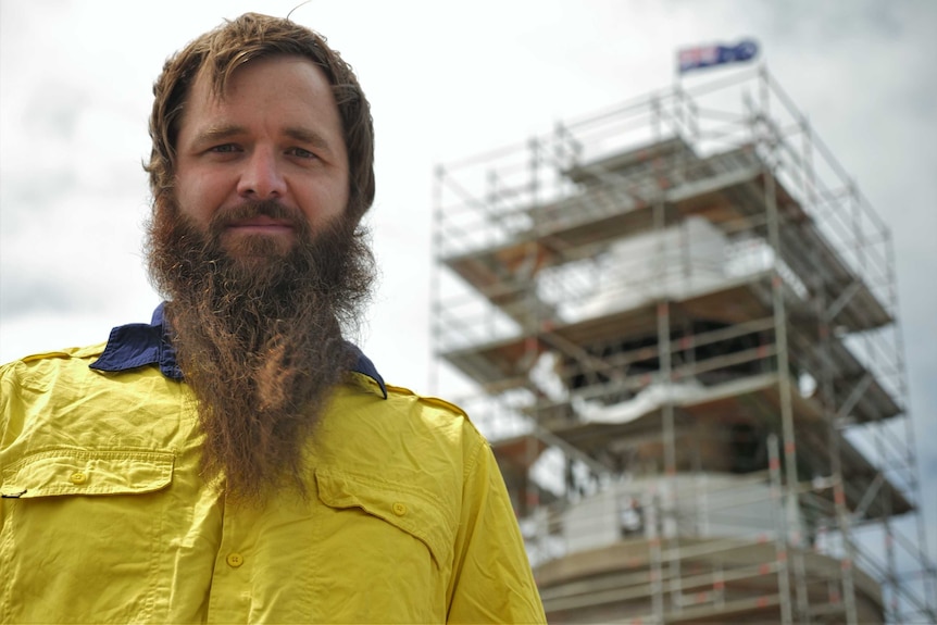 A man wearing a hi-vis shirt with an impressive beard stands in front of a lighthouse on an island.