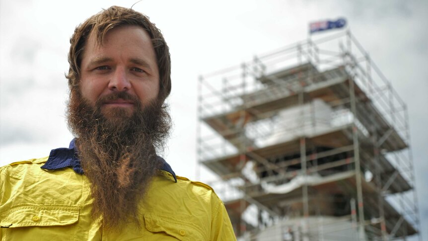 A man wearing a hi-vis shirt with an impressive beard stands in front of a lighthouse on an island.