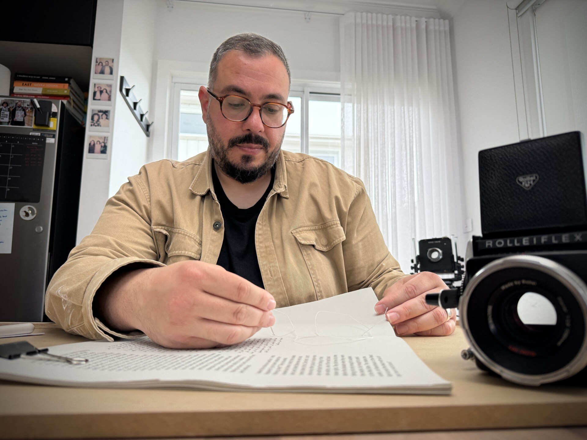 A man in a light brown jacket and black top sits at a desk with an old camera in front of him.