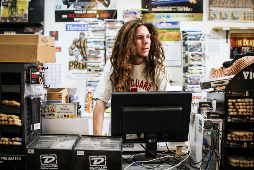 Reece Newling with long red wavy hair standing behind the counter at the local video store he works in.