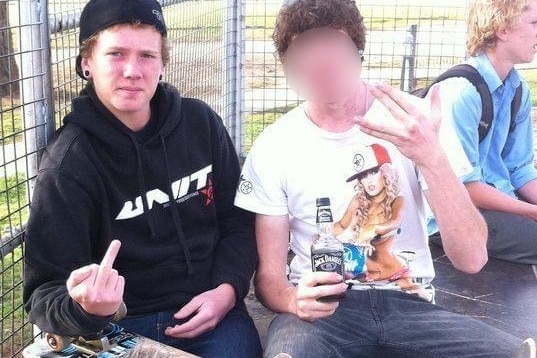 A teenager holding a skateboard flips off the camera. Another teenager sitting next to him is drinking.