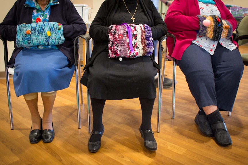 Three elderly women sit in chairs with their hands tucked inside knitted fiddle muffs.