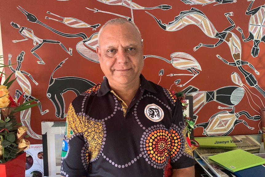 John Berto sits behind his desk with an Aboriginal painting behind him.