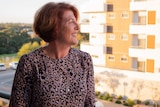 A middle-aged woman with short red hair stands on a balcony looking out to another apartment block