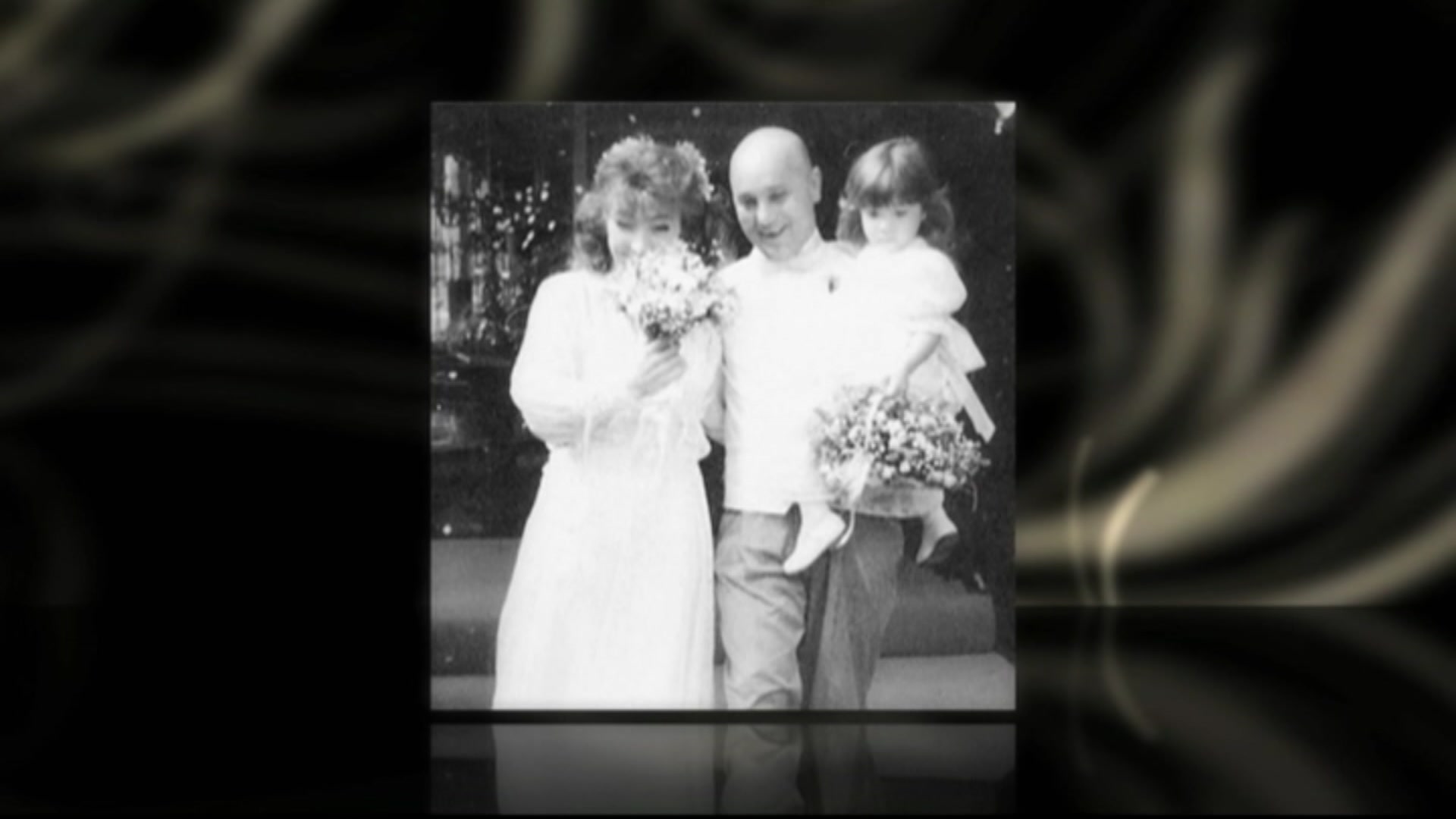 A black and white photo of a woman in a wedding dress with flowers in front of her face and a bald man holding a young girl.