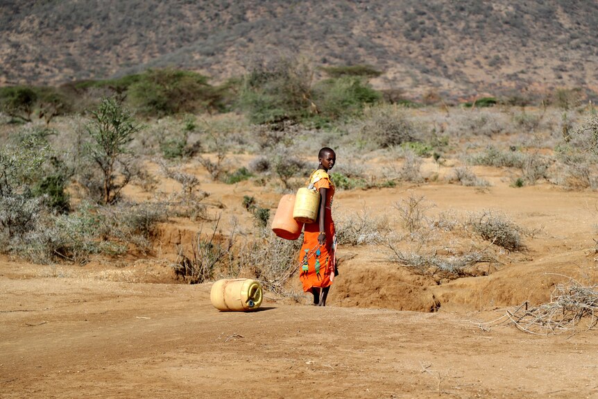 a woman in orange traditional dress with two plastic vessels slung over her shoulder stands in parched scrub