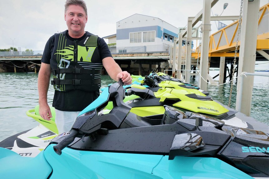Man smiling wearing a buoyancy aid standing on a dock beside a fleet of jetskis.