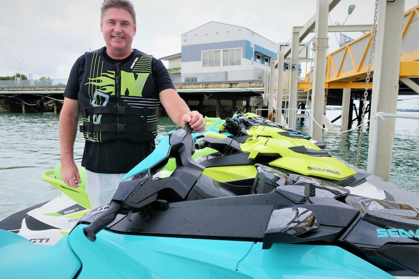 Man smiling wearing a buoyancy aid standing on a dock beside a fleet of jetskis.