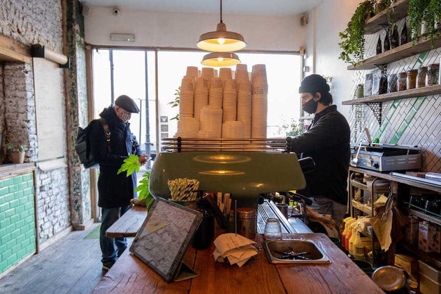 A barista in a mask prepares a takeaway coffee for a customer.