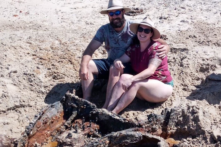 A couple sit in a rusty car body buried in the sand.