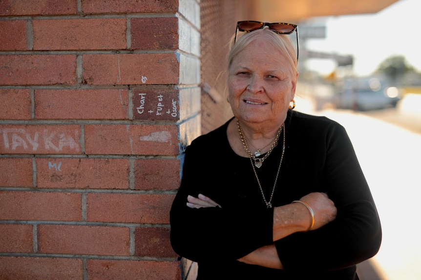 A woman wearing a black top and necklaces folds her arms and leans on a brick wall.