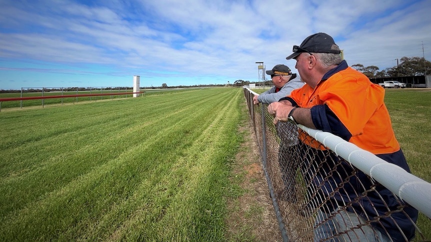 Two men standing on a horse racing track looking at green grass and blue skies.