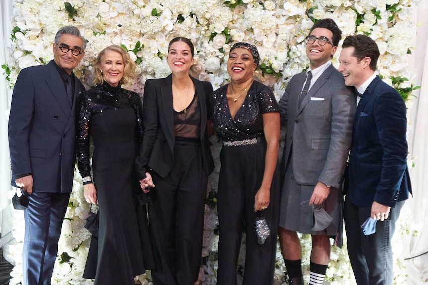 Schitt's Creek cast stands in front of a flower wall.