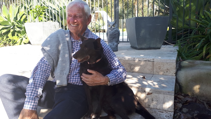 A man with white hair and check shirt sits with his arm around a brown Kelpie.