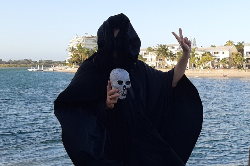 A man in a black cloak, holding a fake skull and giving the peace sign