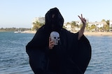 A man in a black cloak, holding a fake skull and giving the peace sign