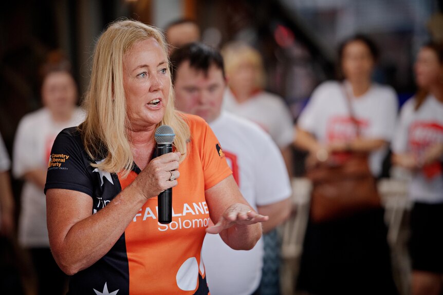 a woman wearing an orange tee shirt holds a microphone and is addressing a crowd of people.