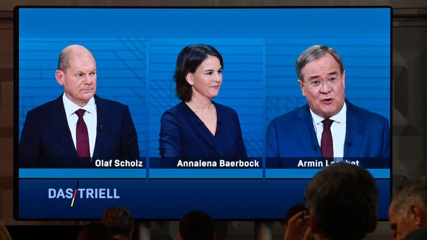 large television screen displaying three main candidates in the german elections, two turning to look at armin laschet.