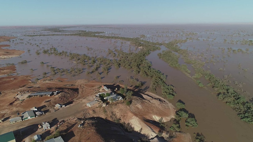 A building on a hill overlooks an expanse of brown floodwater dotted with trees.