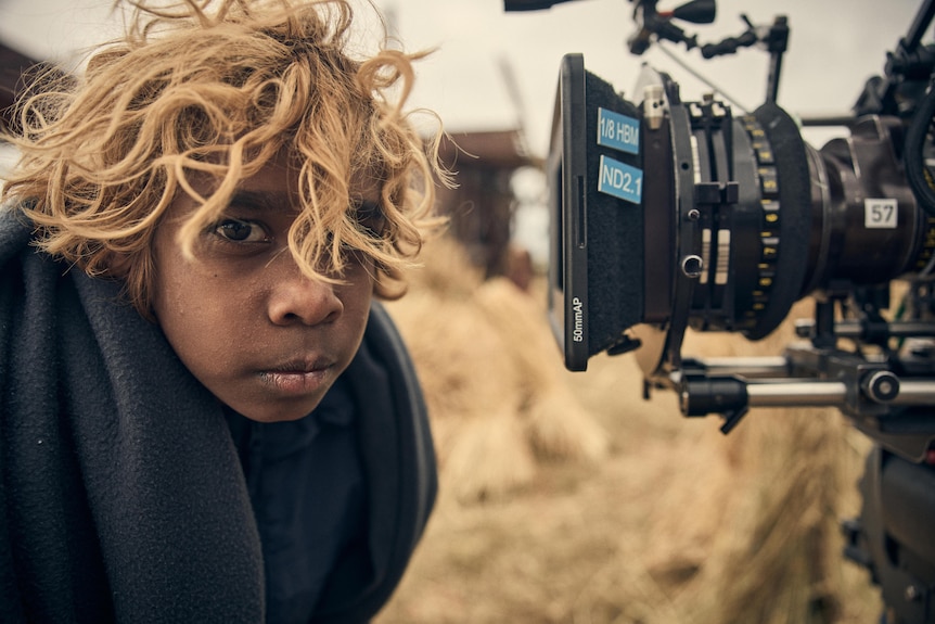 A young Indigenous boy with curly blonde hair looks sideways at the camera, away from the lens of a film camera.