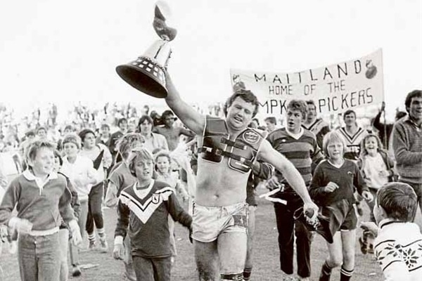 a black and white photo of a man in the 50's holding a rugby league trophy in front of a crowd