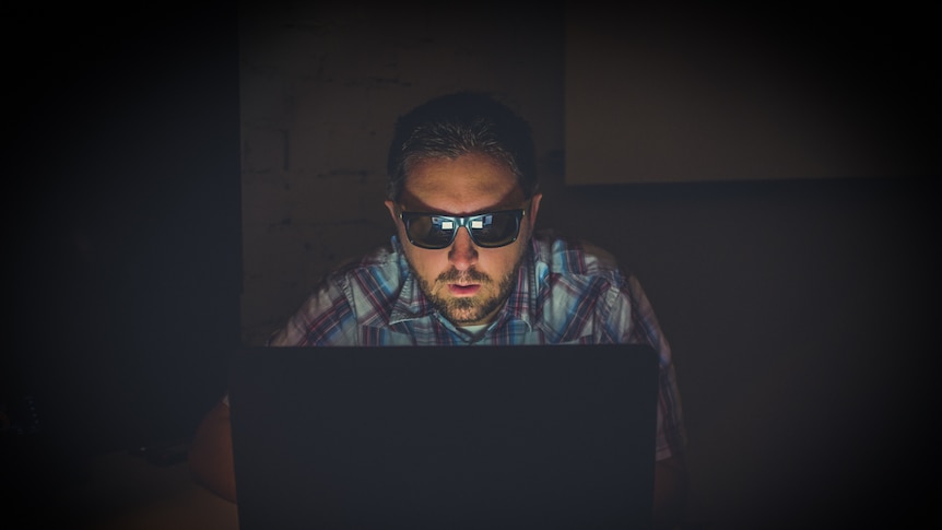 A man sitting in the dark using a laptop wearing black sunglasses.