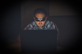 A man sitting in the dark using a laptop wearing black sunglasses.
