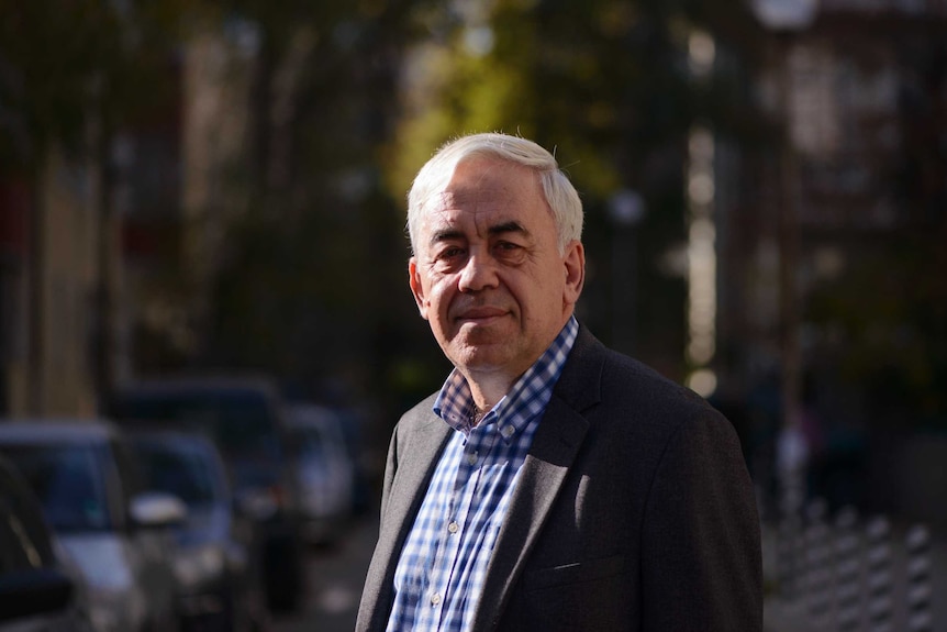 A man in a blue check shirt and wearing a jacket stands in a street.