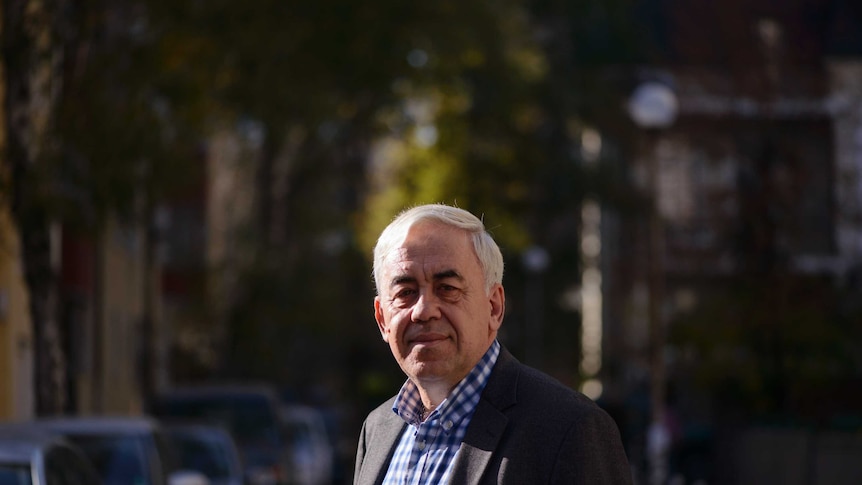 A man in a blue check shirt and wearing a jacket stands in a street.