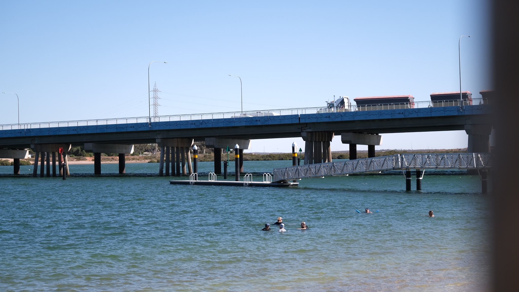 Uma imagem ampla de uma praia com uma ponte sobre ela e pessoas nadando. 