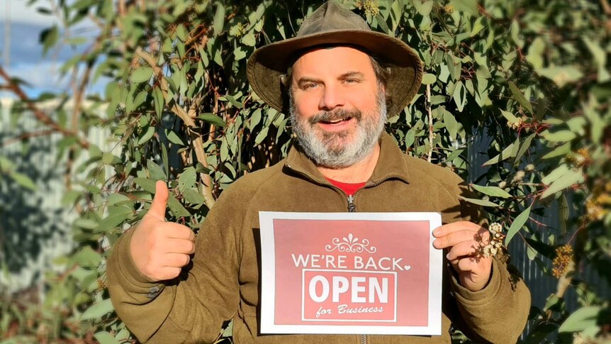 A man stands in front of eucalyptus tree and holds a sign that says 'We're back and open for business' while doing a thumbs up.