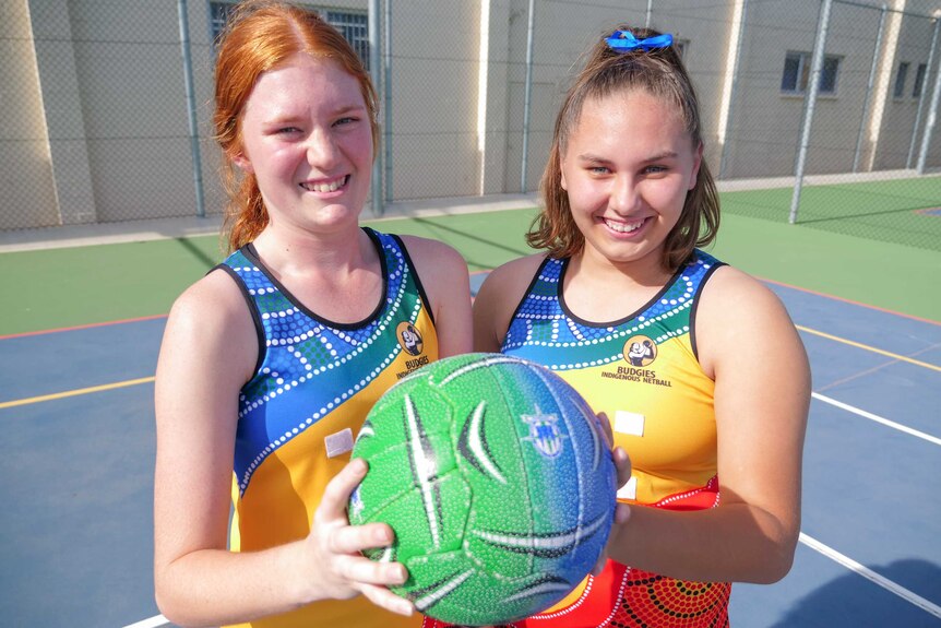 Poppy and Mercedes in their colour Australian Indigenous Budgies uniform holding a netball and smiling.