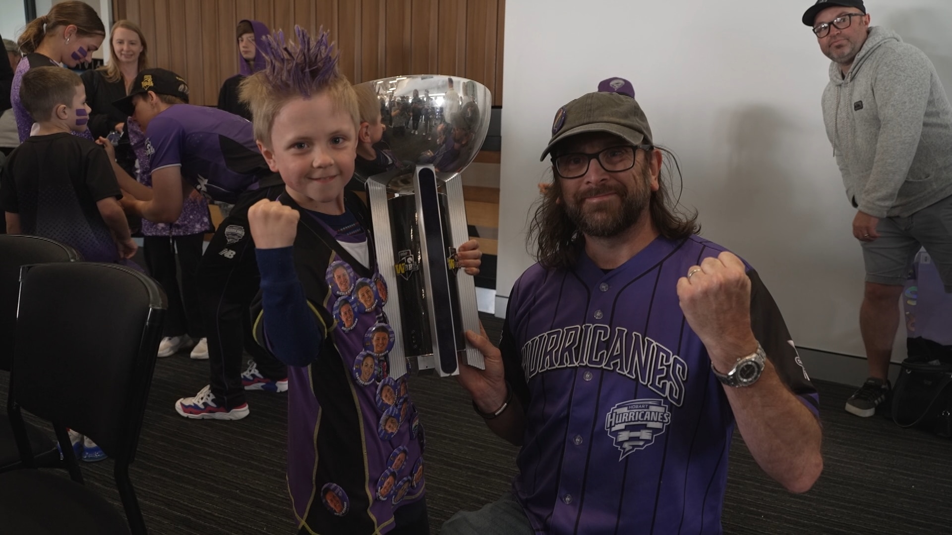 Un niño con cabello rubio con puntas moradas y un hombre sostienen el trofeo de los Huracanes y aprietan los puños.