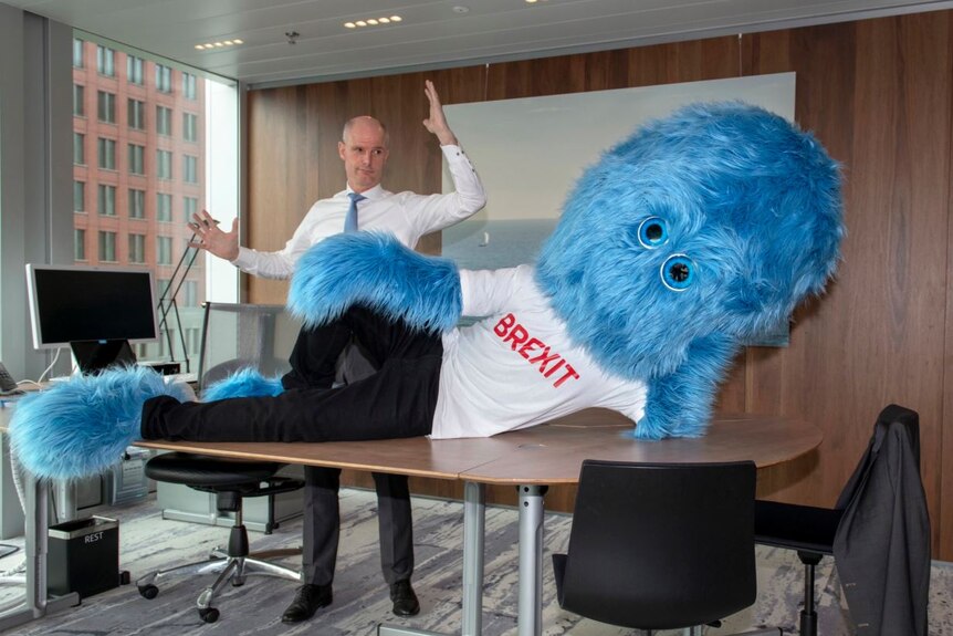 A corporate boardroom with warm wood panelling houses a desk with a furry blue monster reclining on it, with a man behind.
