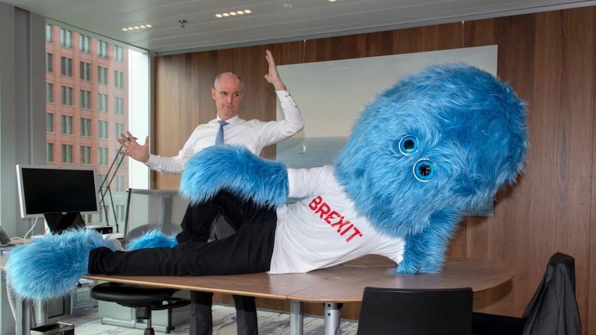 A corporate boardroom with warm wood panelling houses a desk with a furry blue monster reclining on it, with a man behind.