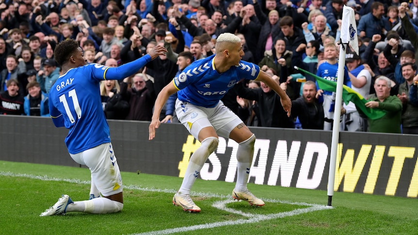 A blonde-haired Everton footballer crouches near the corner flag as a teammate slides in on his knees.