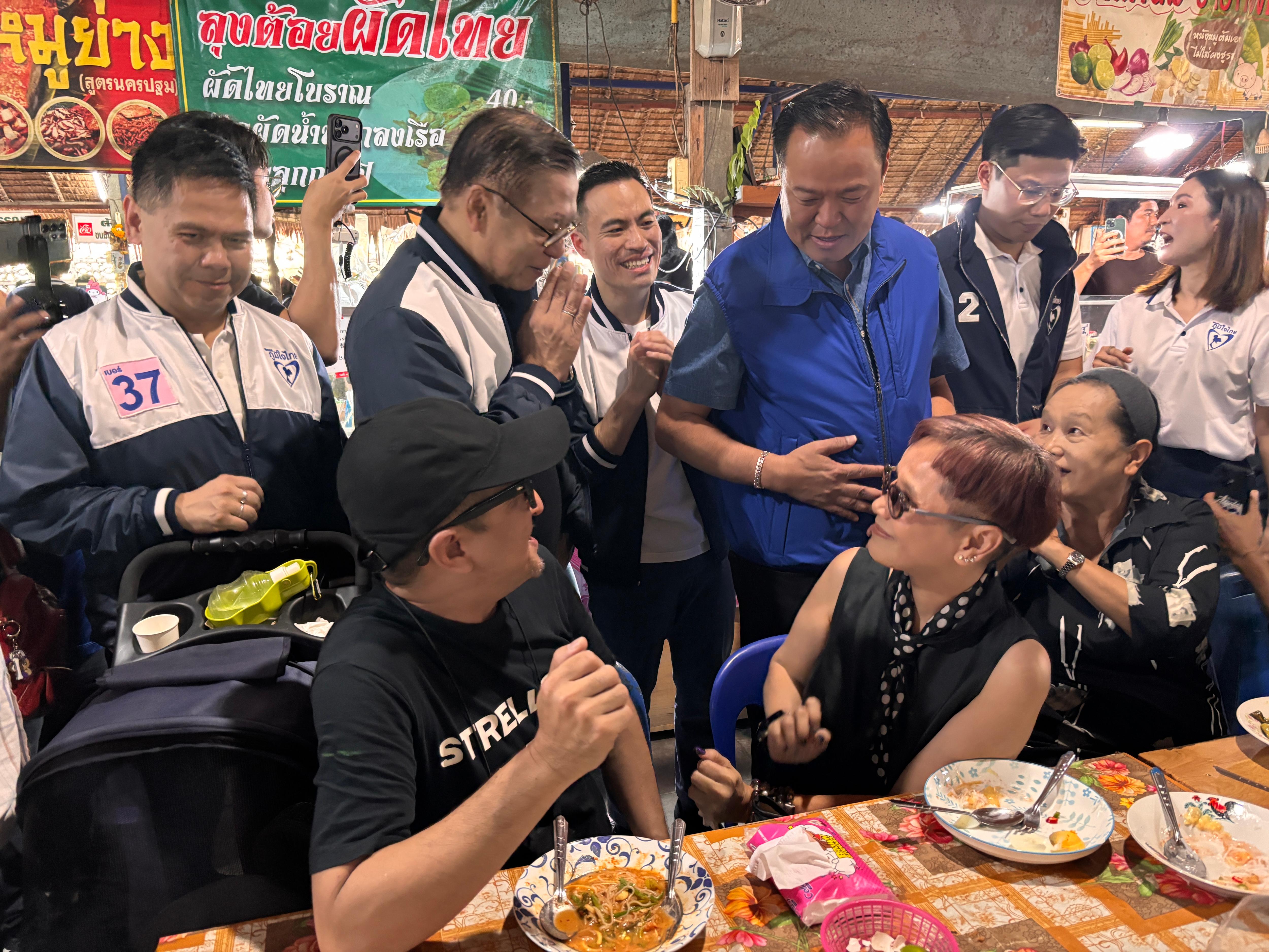 Un hombre con un chaleco azul está detrás y habla con los clientes sentados en una mesa en un mercado.