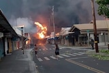 People stand on a street and watch a fire raging in the background