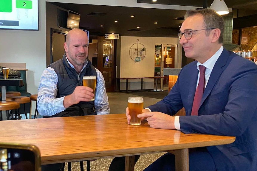 Two men sit at a tall table drinking a beer while being filmed