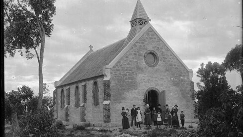 A small group of men, women and children stand in front of the church which has bush and trees around it